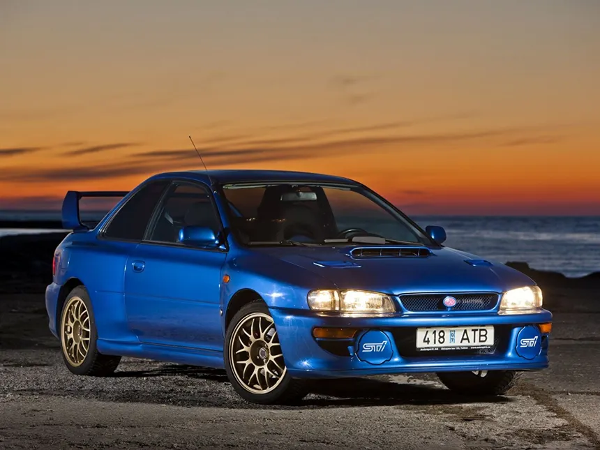 Subaru Impreza 22B STi en azul Sonic Blue Mica estacionado en un tramo de rally de montaña con camino de grava y bosque al fondo durante la hora dorada
