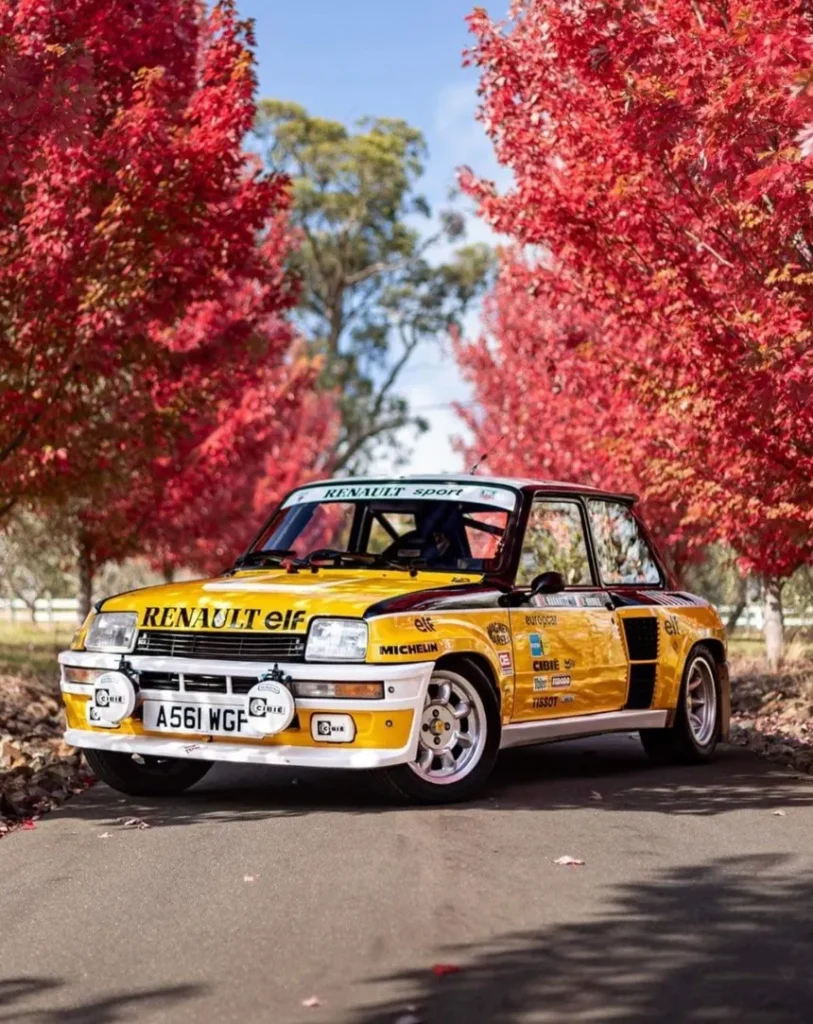 White Renault 5 Turbo 2 with widened arches and lateral air intakes, viewed from three-quarter rear angle on a mountain road