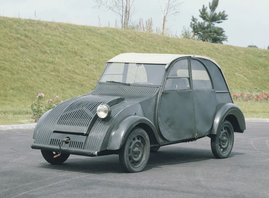 Citroën 2CV gris claro y Citroën AK400 azul aparcadas en un patio rural francés, vistas tres cuartos delanteras, mostrando claramente el morro común y las diferentes carrocerías
