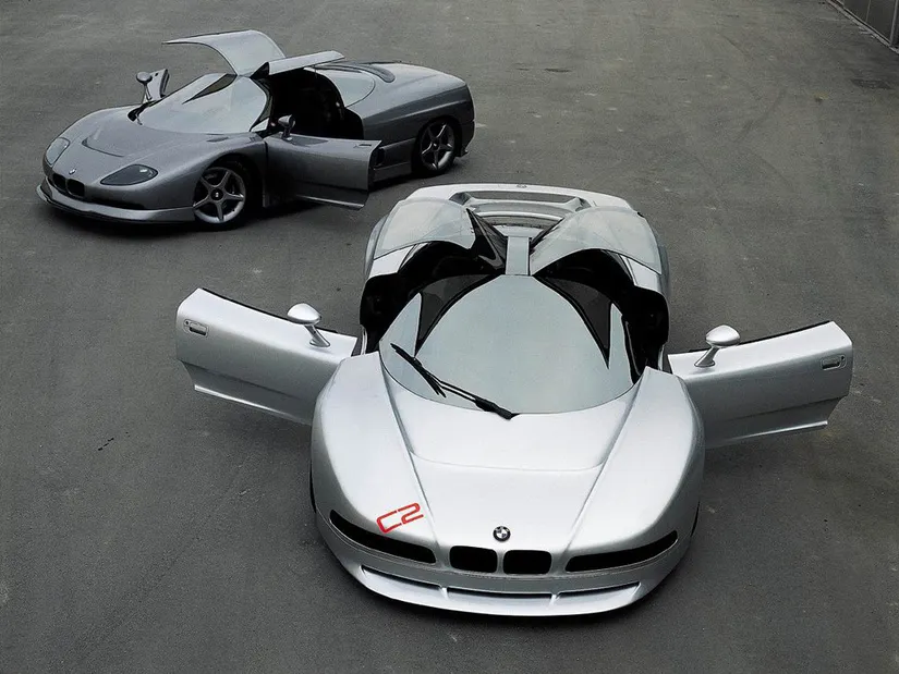 Nazca C2 coupé 3/4 frontal con ventanas semi-gullwing abiertas, sobre fondo neutro tipo salón. 