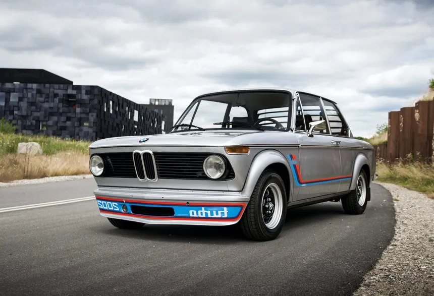 BMW 2002 turbo 1973 en color blanco Chamonix con franjas Motorsport azul-violeta-rojo, arcos plásticos atornillados y calcomanía especular "OBRUT 2002" en el spoiler delantero