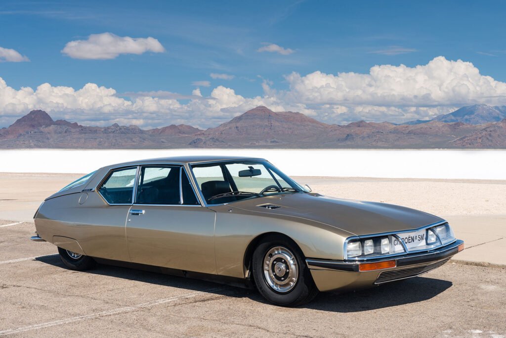 1972 metallic grey Citroën SM, three-quarter front view on a French mountain road, swivelling headlights deployed, glass-covered grille showing six visible lamps
